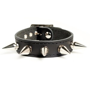 Può includere: Braccialetto in pelle nera con punte argentate. Il braccialetto è largo circa 5 cm e ha cinque punte.