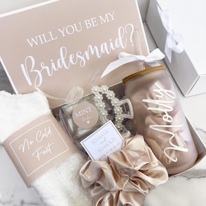 May include: A gift box with the text "WILL YOU BE MY Bridesmaid?" contains a personalized tumbler, scrunchies, a hair clip, mints, and socks. The tumbler is light pink with the name "Molly" in white script. The socks have the text "No Cold Feet".