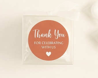 May include: A round, orange thank you favor tag with white text that reads "Thank You for Celebrating with Us" and a small white heart. The tag is in a clear plastic box with a white ribbon.