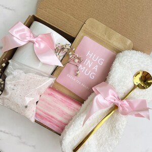 May include: A gift box filled with a white mug, pink bow, bath salts, soap, a gold spoon, and a packet of green tea. The pink card reads "HUG IN A MUG". The box is tied with a pink satin ribbon.