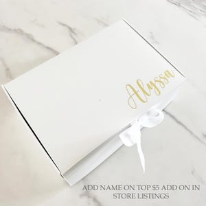May include: A white gift box with a gold script name "Alyssa" on the top. A white satin ribbon is tied in a bow on the front. The text "ADD NAME ON TOP $5 ADD ON IN STORE LISTINGS" is at the bottom.