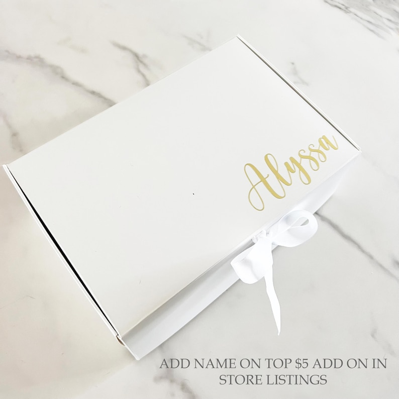 May include: A white gift box with a gold script name "Alyssa" on the top. A white satin ribbon is tied in a bow on the front. The text "ADD NAME ON TOP $5 ADD ON IN STORE LISTINGS" is at the bottom.
