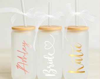 May include: Three frosted glass tumblers with bamboo lids and clear straws. The tumblers are personalized with the names "Ashley", "Bride", and "Katie" in different colors.