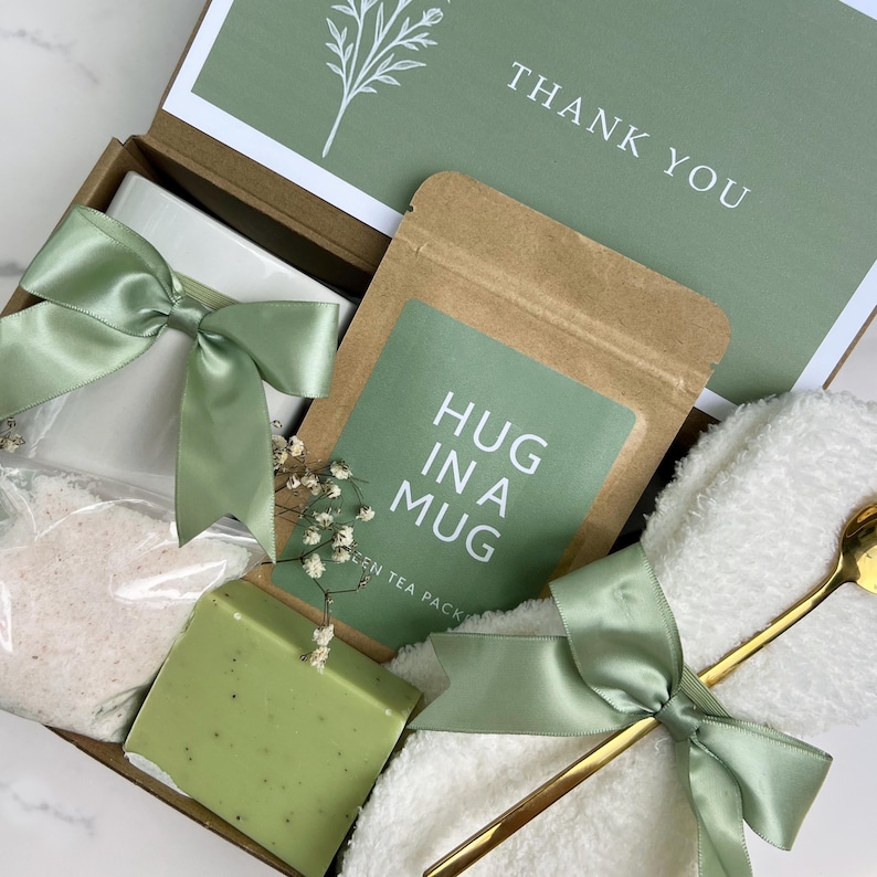 May include: A gift box with a white mug tied with a sage green ribbon, a soap bar, a tea packet labeled "HUG IN A MUG", a gold spoon, and a "THANK YOU" card. The box is filled with a white towel and bath salts.
