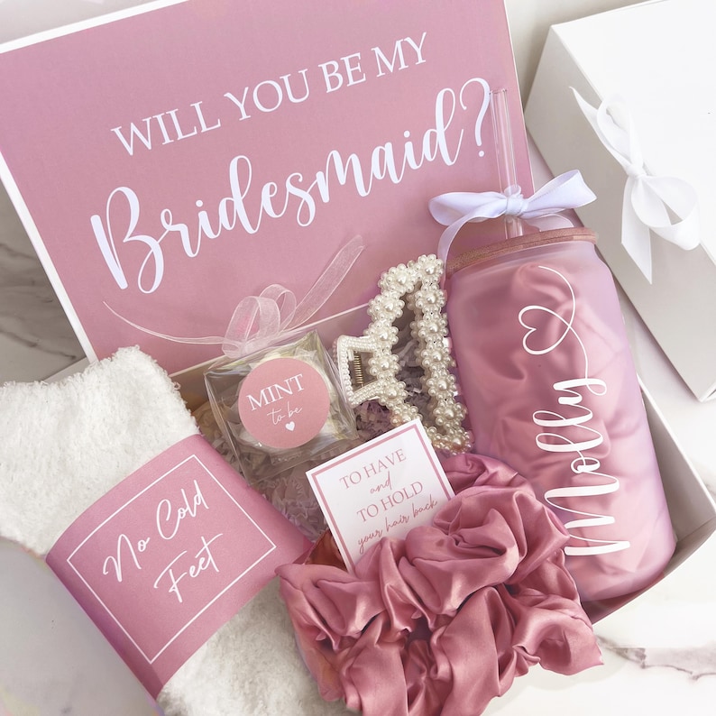 May include: A pink gift box with the text "WILL YOU BE MY Bridesmaid?" contains a tumbler, hair accessories, socks, and a small mint. The tumbler has a straw and the name "Molly" on it.