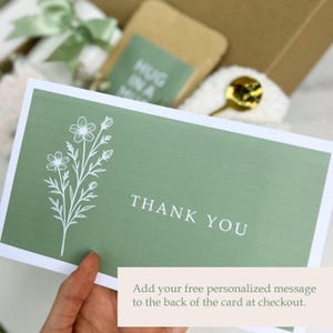 May include: A sage green card with white floral illustrations and the words "THANK YOU" in white. The card is held in a hand, with a gift box and other items visible in the background. Text at the bottom reads "Add your free personalized message to the back of the card at checkout."
