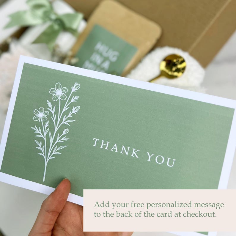 May include: A sage green card with white floral illustrations and the words "THANK YOU" in white. The card is held in a hand, with a gift box and other items visible in the background. Text at the bottom reads "Add your free personalized message to the back of the card at checkout."