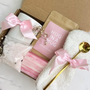 May include: A gift box filled with a white mug, pink bow, bath salts, soap, a gold spoon, and a packet of green tea. The pink card reads "HUG IN A MUG". The box is tied with a pink satin ribbon.