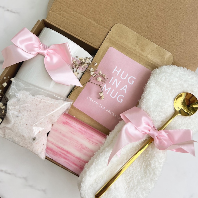 May include: A gift box filled with a white mug, pink bow, bath salts, soap, a gold spoon, and a packet of green tea. The pink card reads "HUG IN A MUG". The box is tied with a pink satin ribbon.