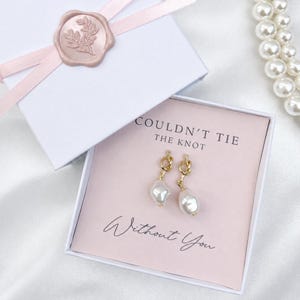 May include: A pair of gold-toned knot earrings with dangling white baroque pearls presented in a white gift box. The box features a pink ribbon with a floral wax seal and a card printed with the text "COULDN'T TIE THE KNOT Without You."