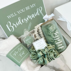 May include: A gift box with a green and white theme, containing a tumbler, scrunchie, socks, and other items. The box lid reads "WILL YOU BE MY Bridesmaid" in white script. The tumbler is personalized with a name in white script. The box also includes a card that reads "TO HAVE AND TO HOLD your hair back."