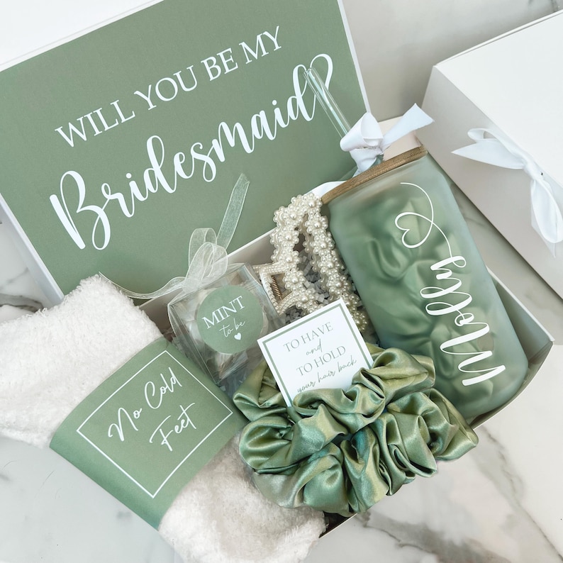 May include: A gift box with a green and white theme, containing a tumbler, scrunchie, socks, and other items. The box lid reads "WILL YOU BE MY Bridesmaid" in white script. The tumbler is personalized with a name in white script. The box also includes a card that reads "TO HAVE AND TO HOLD your hair back."