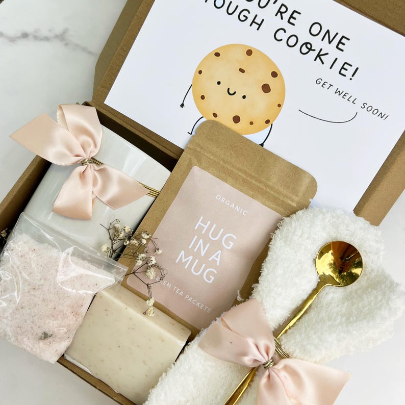 May include: A get-well gift box with a white mug, bath salts, soap, tea packets, and a gold spoon. The box includes a card with a cartoon cookie and the text "You're one tough cookie! Get well soon!" tied with a pink ribbon.