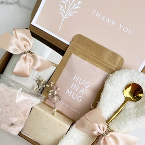 May include: A gift box with a white mug tied with a peach satin bow, a packet of tea labeled "HUG IN A MUG", a bar of soap, bath salts, and a gold spoon. The box also contains a card that says "THANK YOU".