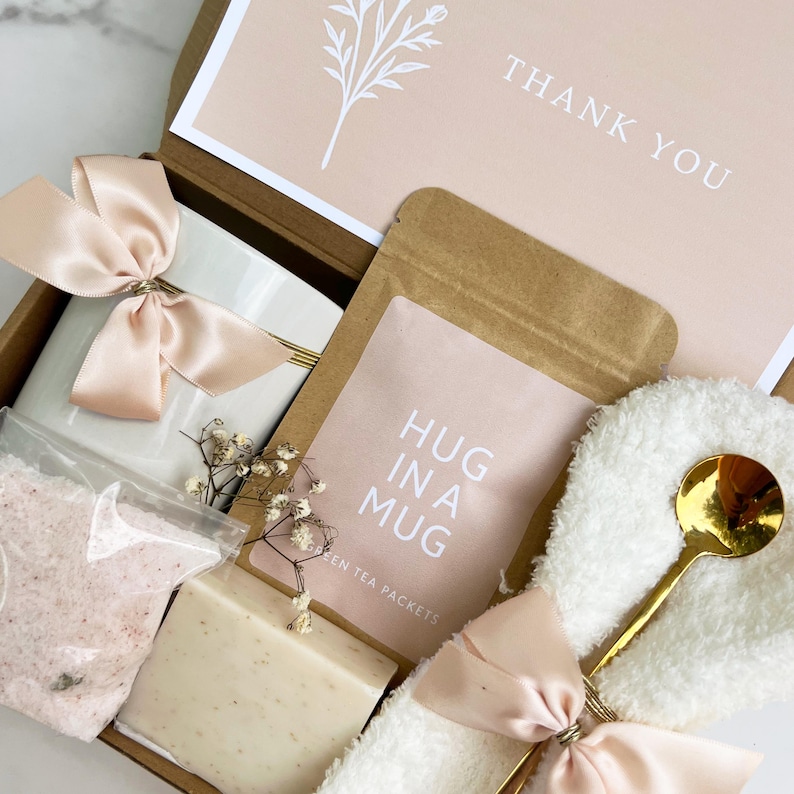 May include: A gift box with a white mug tied with a peach satin bow, a packet of tea labeled "HUG IN A MUG", a bar of soap, bath salts, and a gold spoon. The box also contains a card that says "THANK YOU".