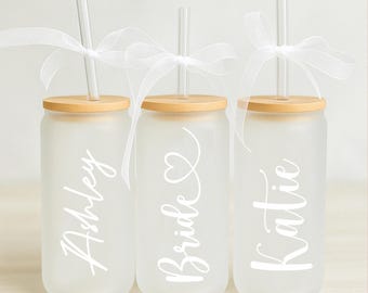 May include: Three frosted glass tumblers with bamboo lids and clear straws. The tumblers are personalized with the names "Ashley", "Bride", and "Katie" in white script.