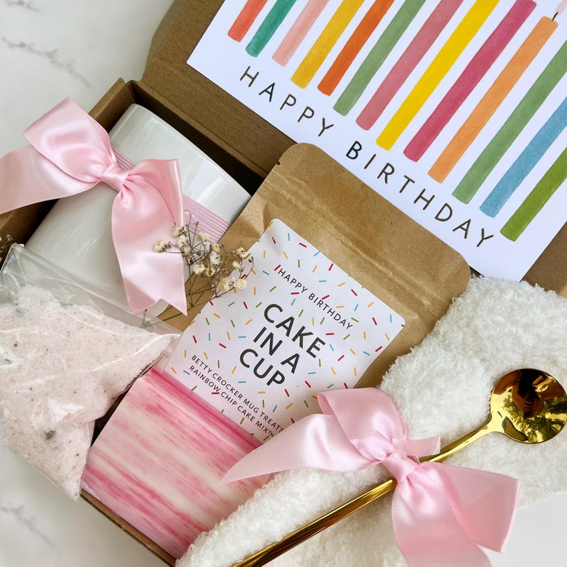 May include: A birthday gift box containing a white mug, a cake mix sachet, a gold spoon, and a card with "Happy Birthday." The box is tied with a pink ribbon. The card features colourful vertical stripes.