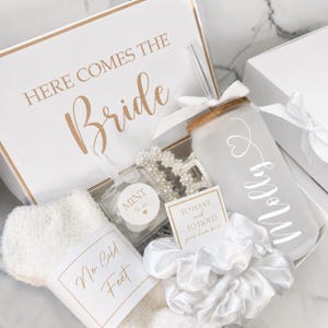 May include: A gift box labeled HERE COMES THE Bride containing a frosted glass tumbler with a straw and the name Molly, a pearl-embellished hair claw, a white satin scrunchie with a card reading TO HAVE and TO HOLD your hair back, and white fuzzy socks with a tag reading No Cold Feet and a mint tin.