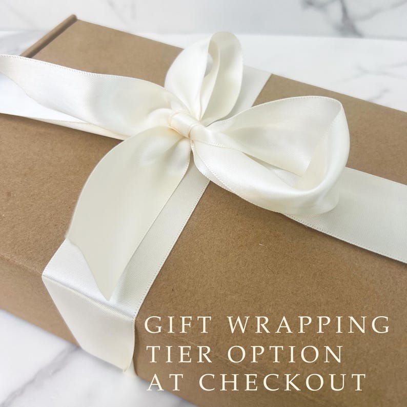 May include: A brown gift box tied with a cream satin ribbon and bow. The text "GIFT WRAPPING TIER OPTION AT CHECKOUT" is printed on the box. The box is presented on a white marble surface.