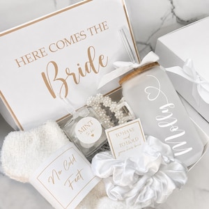 May include: A gift box labeled HERE COMES THE Bride containing a frosted glass tumbler with a straw and the name Molly, a pearl-embellished hair claw, a white satin scrunchie with a card reading TO HAVE and TO HOLD your hair back, and white fuzzy socks with a tag reading No Cold Feet and a mint tin.