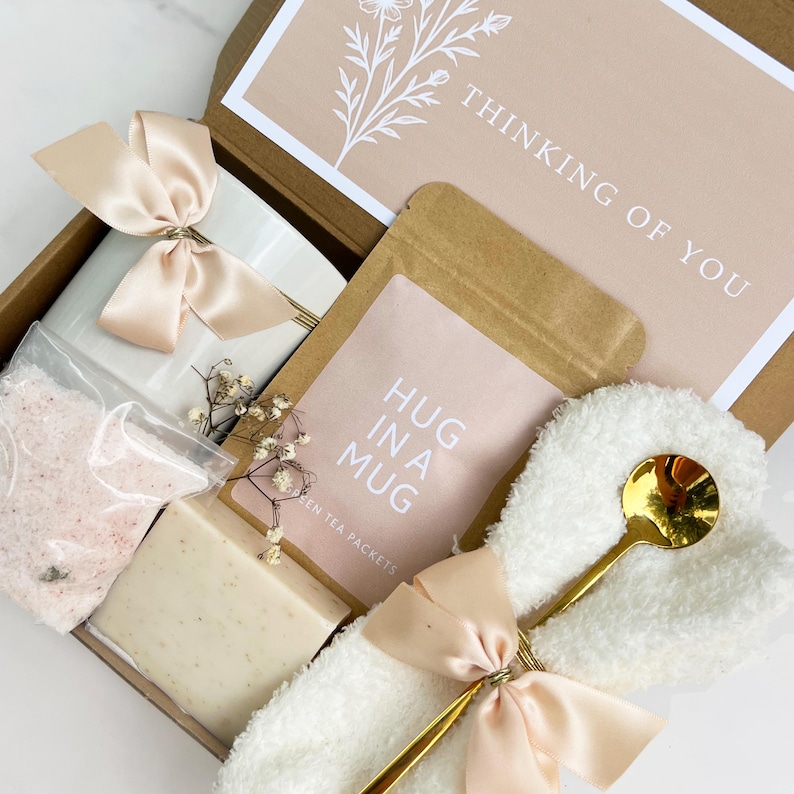 May include: A gift box filled with self-care items. Includes a white mug with a bow, a bag of bath salts, a bar of soap, a tea packet, a washcloth with a gold spoon, and a card that says "Thinking of You."