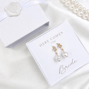 May include: A pair of gold-toned knot earrings with dangling white teardrop pearls, presented in a white square gift box. The box lid features the printed text "HERE COMES THE Bride" in gold lettering, accompanied by a white ribbon and wax seal.