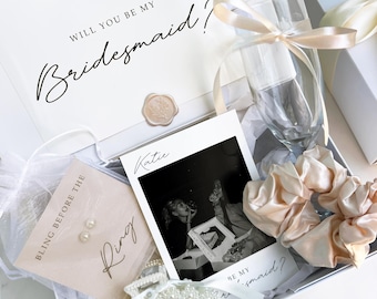 May include: A white gift box containing a card with the text Will You Be My Bridesmaid, a clear champagne flute with a champagne-colored ribbon, a pair of pearl stud earrings on a card labeled Bling Before The Ring, a photo of Katie, and a pearl hair clip.
