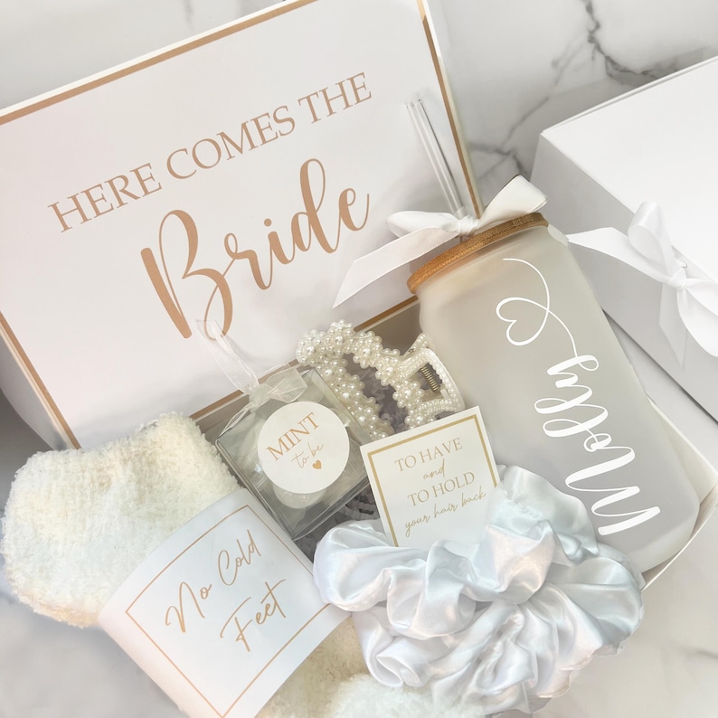 May include: A white gift box with gold trim and the text "HERE COMES THE Bride" contains a frosted glass tumbler with a straw, a pearl bracelet, a scrunchie, and other bridal accessories. The box also includes a pair of white socks labeled "No Cold Feet".