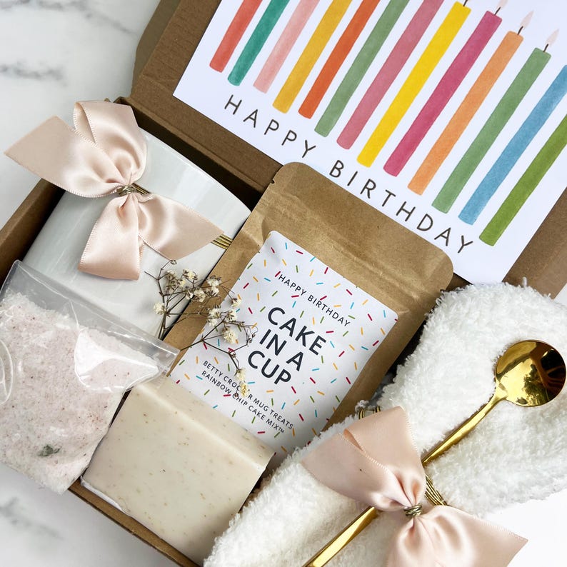 May include: A birthday gift box containing a white mug, bath salts, soap, a cake mix sachet, a gold spoon, and a fluffy white washcloth. The box includes a card with colourful candles and the words "Happy Birthday."