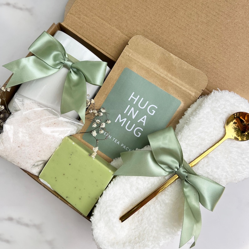 May include: A gift box filled with self-care items. The box contains a white mug, bath salts, a green soap bar, a tea packet with the text "HUG IN A MUG", a white washcloth, and a gold spoon. All items are tied with sage green satin bows.