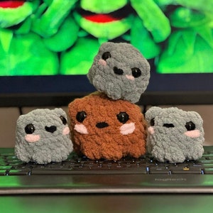May include: Three small crocheted plush toys in the shape of cubes. The top toy is gray, the middle toy is brown, and the bottom toy is gray. The toys have embroidered eyes and pink cheeks. The toys are sitting on a black keyboard.