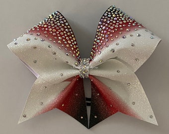 May include: A cheerleading bow with a white, red, and black ombre design. The bow is embellished with sparkling rhinestones and a rhinestone-studded center piece. The bow is made of a glittery material.