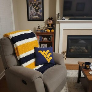 May include: A chunky knit throw blanket featuring horizontal stripes in navy blue, white, and gold draped over a gray recliner. A square navy blue throw pillow with a large gold West Virginia University logo is placed on the seat of the recliner.
