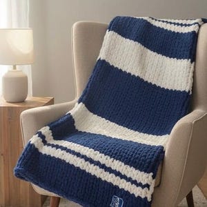 May include: A chunky knit throw blanket featuring wide horizontal stripes in navy blue and cream white. The blanket has a thick, textured yarn construction and includes a small rectangular patch with a blue and white logo on the bottom corner.