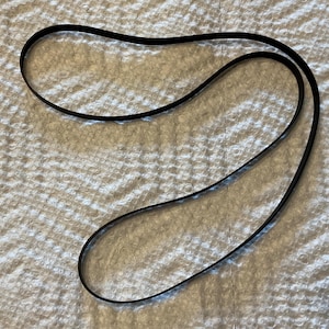 May include: A thin, black, flexible rubber drive belt with a smooth surface and a continuous loop shape. The belt is pliable and maintains a curved, irregular oval form, showing a uniform width and a flat, matte black finish throughout its length.