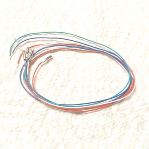May include: A collection of thin, flexible electrical wires in red, white, blue, and green colors. Each wire features a small, cylindrical metal connector crimped onto one end, while the opposite ends are stripped to reveal exposed conductive metal.