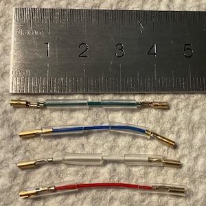 May include: Four electrical wire connectors with gold-colored metal terminals at each end and clear plastic insulation sleeves. The wires are green, blue, white, and red. A metal ruler shows a scale in millimeters for size reference.