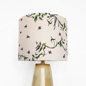 Puede incluir: Una pantalla de lámpara de color crema con un patrón floral que presenta vides verdes y abejas. La pantalla de la lámpara está sobre una base de lámpara de cerámica beige.