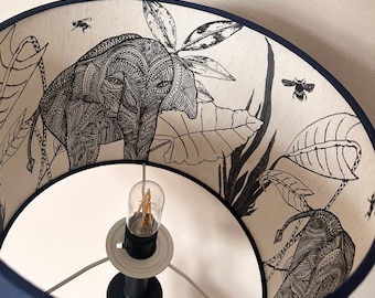 May include: A black and white patterned lampshade with an elephant design. The lampshade is made of fabric and has a navy blue trim. The lampshade is lit with a light bulb.