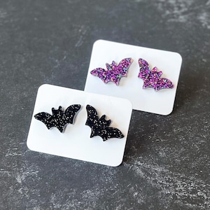 May include: Two pairs of bat-shaped stud earrings displayed on white rectangular cards. One pair features a black base with fine silver glitter, while the other pair features a purple and pink glitter finish. Each bat has outstretched wings.