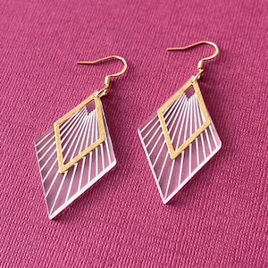 May include: A pair of geometric dangle earrings. Each earring features a clear, diamond-shaped pendant with white linear details and a gold-colored square frame. The earrings hang from gold-colored ear wires, set against a pink background.
