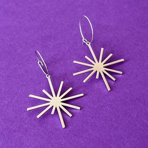 May include: A pair of gold-colored starburst earrings with a simple hook design. The earrings are set against a solid purple background, creating a striking contrast. The starburst design features multiple rays extending from a central point.