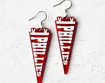 May include: A pair of red acrylic earrings shaped like pennants with the word "PHILLIES" written in white letters.