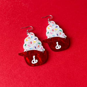 May include: A pair of cupcake-shaped earrings. Each earring features a red base with a white swirl of frosting topped with colorful sprinkles. The earrings have silver-colored ear wires.