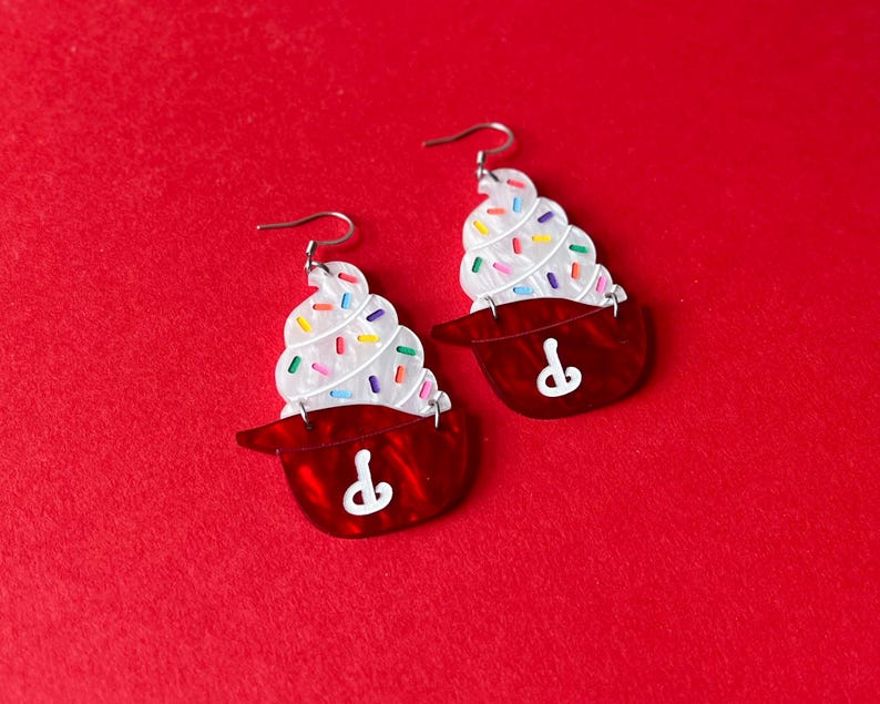 May include: A pair of cupcake-shaped earrings. Each earring features a red base with a white swirl of frosting topped with colorful sprinkles. The earrings have silver-colored ear wires.