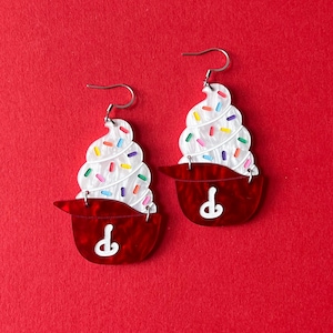May include: A pair of novelty earrings shaped like cupcakes. Each earring features a white swirl of frosting with colorful sprinkles atop a red cupcake liner. The earrings have silver-colored hooks.