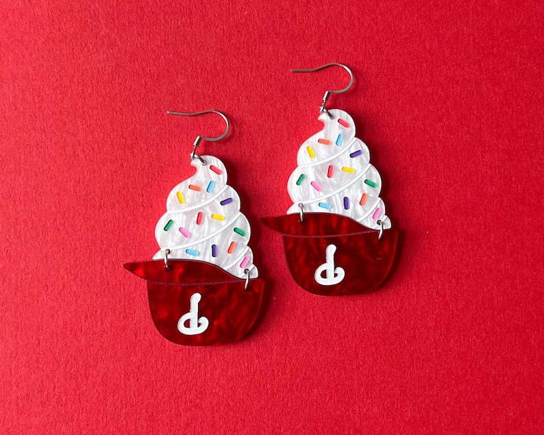 May include: A pair of novelty earrings shaped like cupcakes. Each earring features a white swirl of frosting with colorful sprinkles atop a red cupcake liner. The earrings have silver-colored hooks.