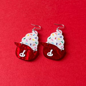 May include: A pair of novelty earrings shaped like cupcakes. Each earring features a red base with a white "P" design and a swirl of white frosting topped with colorful sprinkles. The earrings have silver-colored ear wires.