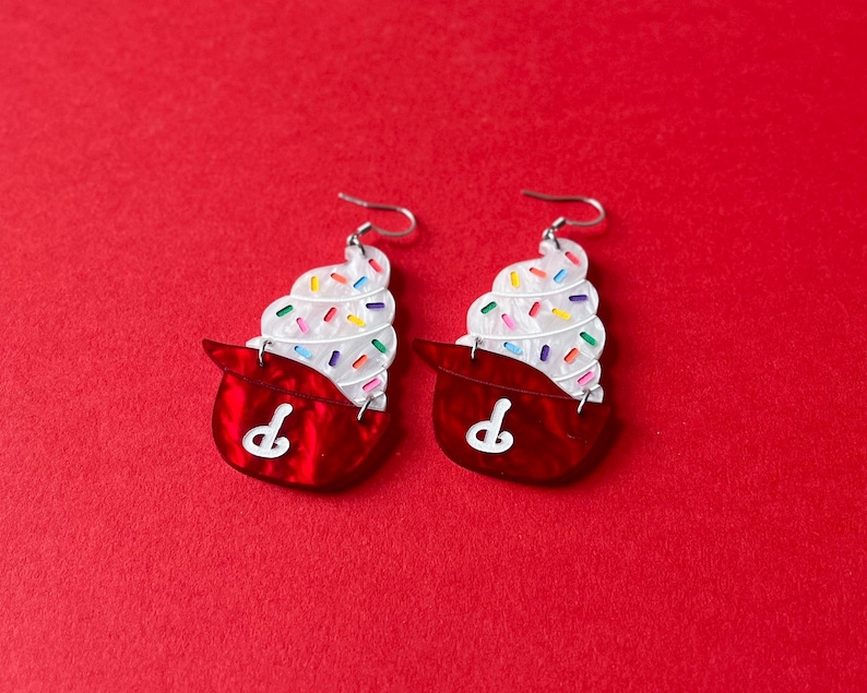 May include: A pair of novelty earrings shaped like cupcakes. Each earring features a red base with a white "P" design and a swirl of white frosting topped with colorful sprinkles. The earrings have silver-colored ear wires.
