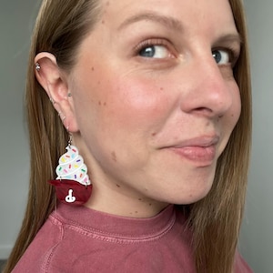 May include: A pair of novelty earrings shaped like cupcakes. The earrings feature a red cupcake holder with a white frosting swirl and colorful sprinkles. The earrings are hanging from silver hooks.